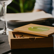 An open bamboo box with a green logo on the lid, containing cutlery placed on a table with a glass and napkin in the background.