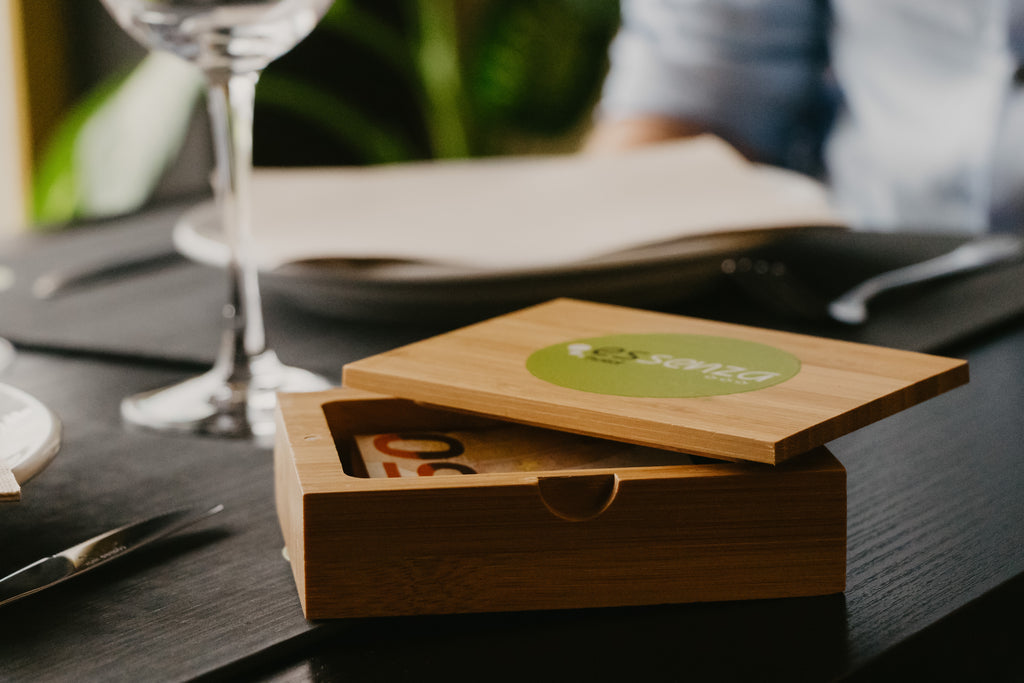 An open bamboo box with a green logo on the lid, containing cutlery placed on a table with a glass and napkin in the background.